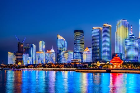 어둠이 내린 해안가 도시의 마천루들이 형형색색의 조명을 밝히고 있으며, 잔잔한 물결 위로 화려한 빛의 반영이 길게 늘어져 환상적인 야경을 선사합니다.