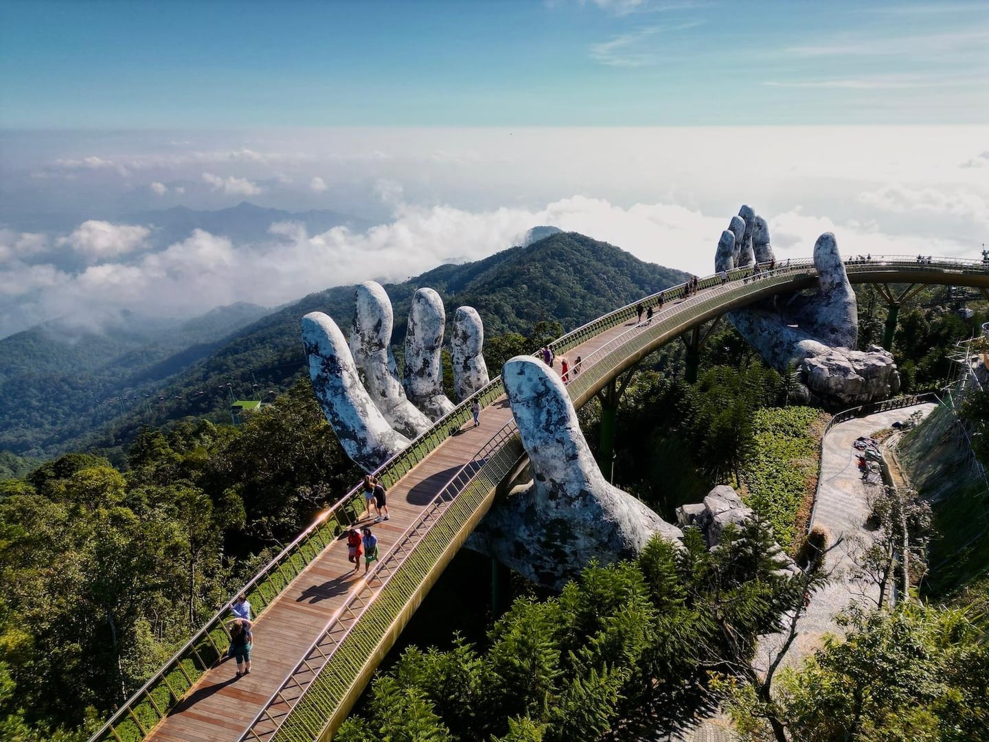 구름이 내려다보이는 높은 산 정상에서 거대한 신의 손 조형물이 황금빛 곡선 다리를 받치고 있는 다낭 바나힐 골든 브릿지의 웅장한 풍경입니다.