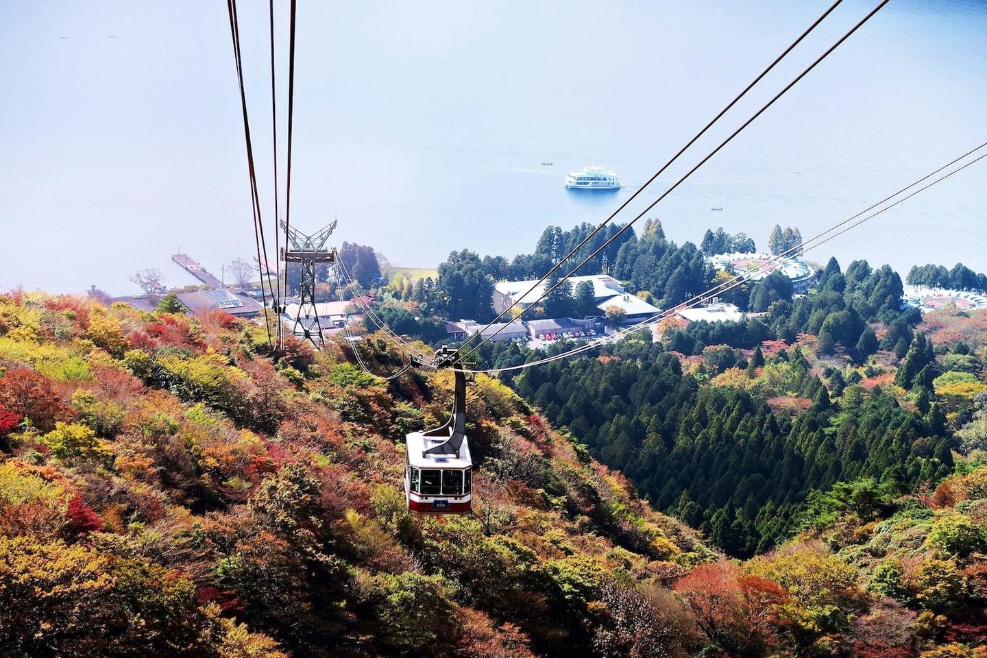 가을 단풍으로 물든 산을 가로지르는 케이블카 너머로 잔잔한 호수와 하얀 유람선이 펼쳐진 아름다운 자연 경관입니다.