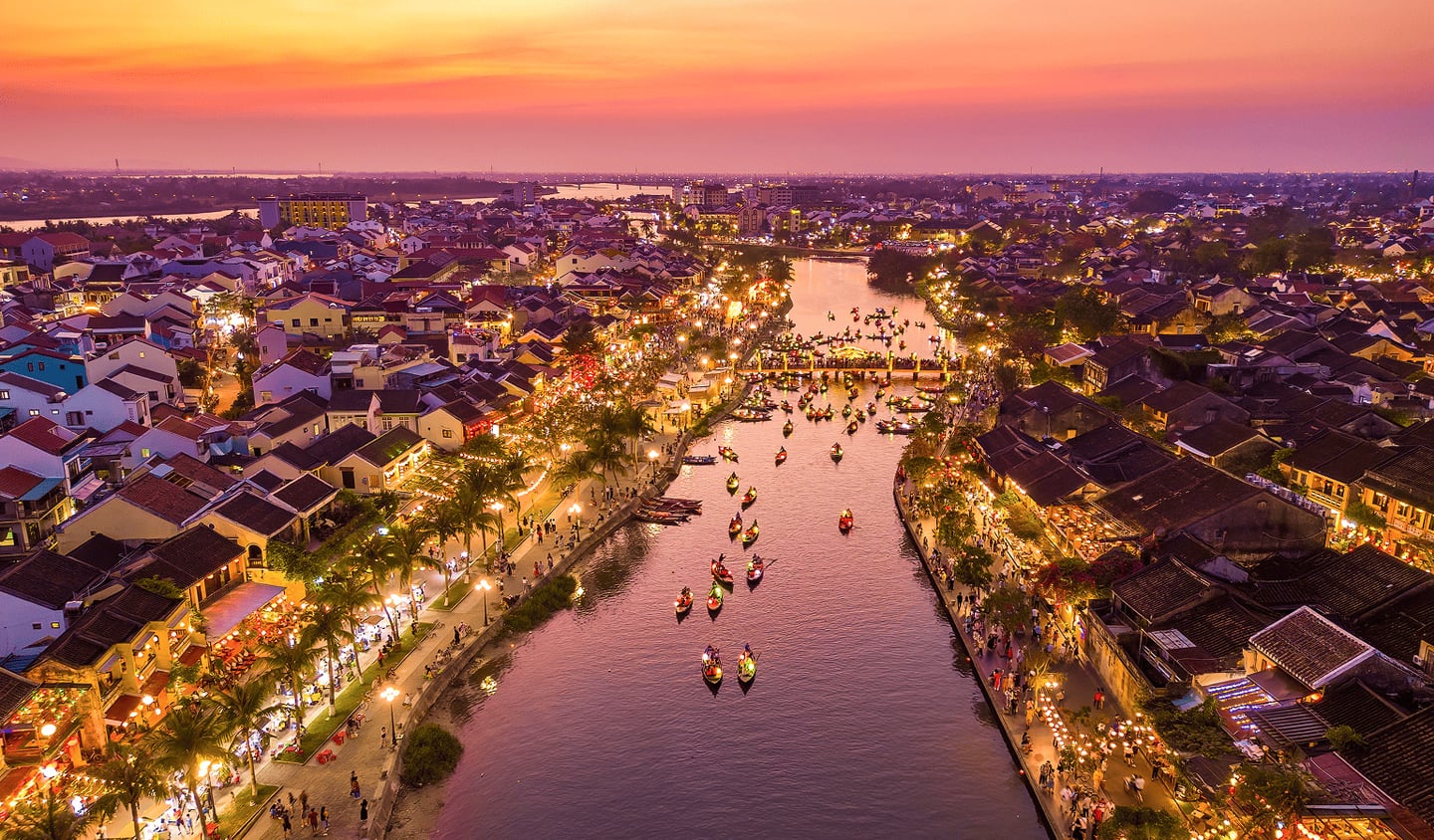 노을이 지는 보랏빛 하늘 아래 수많은 소원 배가 떠 있는 호이안 투본강과 황금빛 조명으로 빛나는 올드타운의 아름다운 야경입니다.