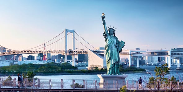 오다이바의 상징인 자유의 여신상 너머로 푸른 바다를 가로지르는 거대한 레인보우 브릿지와 도쿄 타워가 보이는 도심 전경입니다.