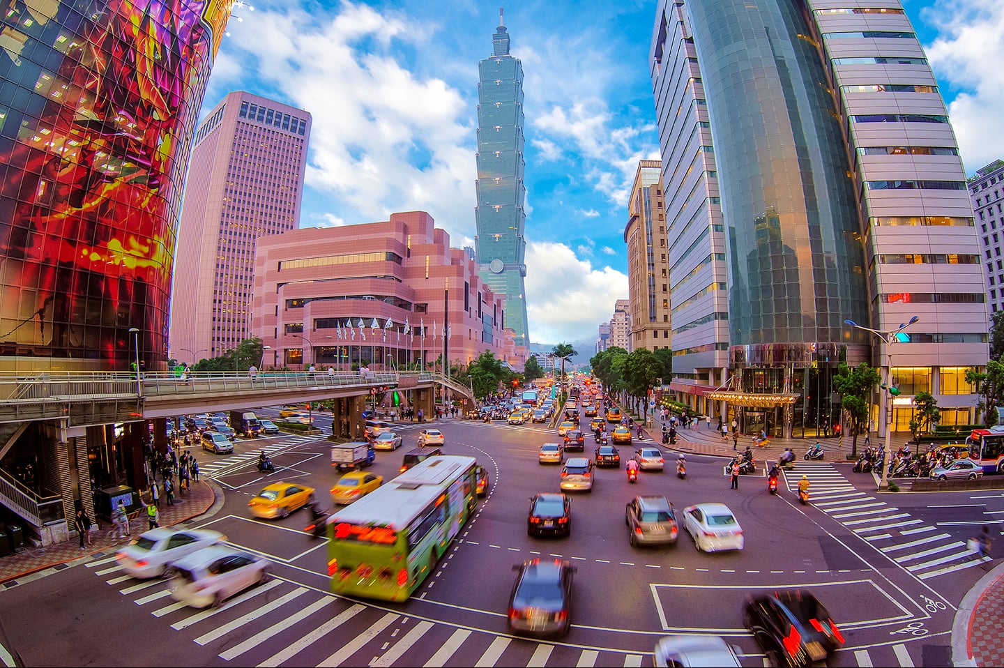푸른 하늘 아래 타이베이 101 타워가 보이는 번화한 도심 교차로에서 수많은 차량이 이동하며 활기찬 도시의 일상을 보여주는 풍경입니다.