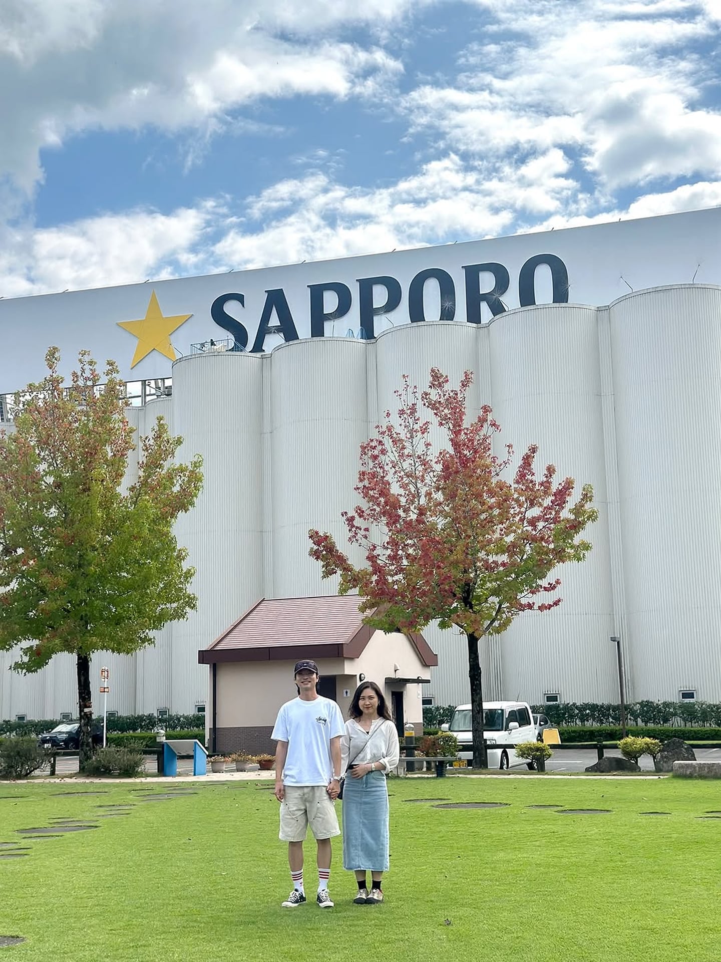 삿포로 맥주 공장의 대형 로고가 보이는 넓은 잔디 광장에서 손을 잡고 서 있는 커플과 붉게 물든 단풍나무가 어우러진 풍경입니다.