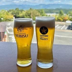탁 트인 야외 전경을 배경으로 시원한 거품이 올라간 에비스와 삿포로 생맥주 잔이 나무 테이블 위에 놓여 있는 모습입니다.