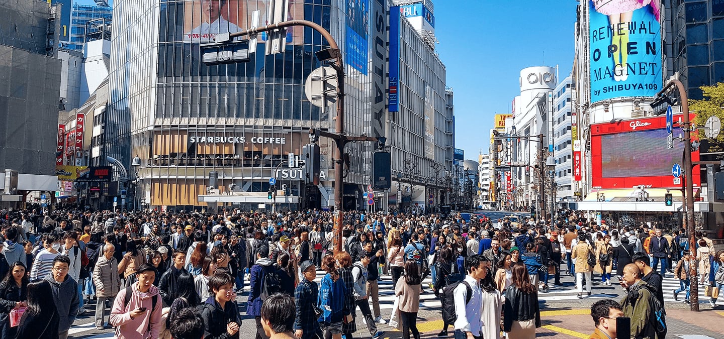맑은 하늘 아래 도쿄 시부야의 번화한 거리에서 수많은 사람들이 횡단보도를 가득 메우며 이동하고 있는 역동적인 도심의 모습입니다.