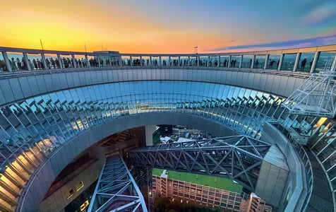 황금빛 노을이 물든 하늘을 배경으로 원형의 공중정원 전망대 난간에 서서 오사카 시내 전경을 즐기는 사람들과 독특한 건축 구조물.