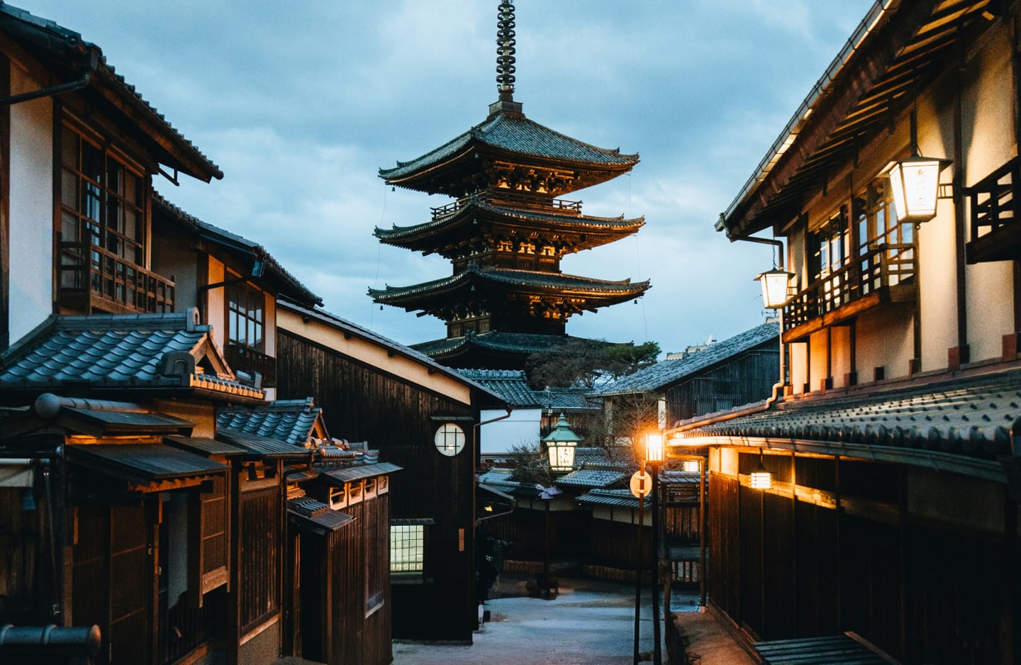 어스름한 저녁 무렵 교토의 전통적인 목조 가옥 거리 끝으로 웅장한 야사카의 탑이 보이고 건물마다 따뜻한 등불이 켜져 아늑한 정취를 풍깁니다.