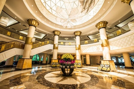 화려한 샹들리에와 금빛 기둥이 어우러진 웅장한 호텔 로비 전경으로, 대리석 바닥 중앙의 꽃 장식과 원형 돔 천장이 럭셔리한 실내 인테리어를 완성합니다.