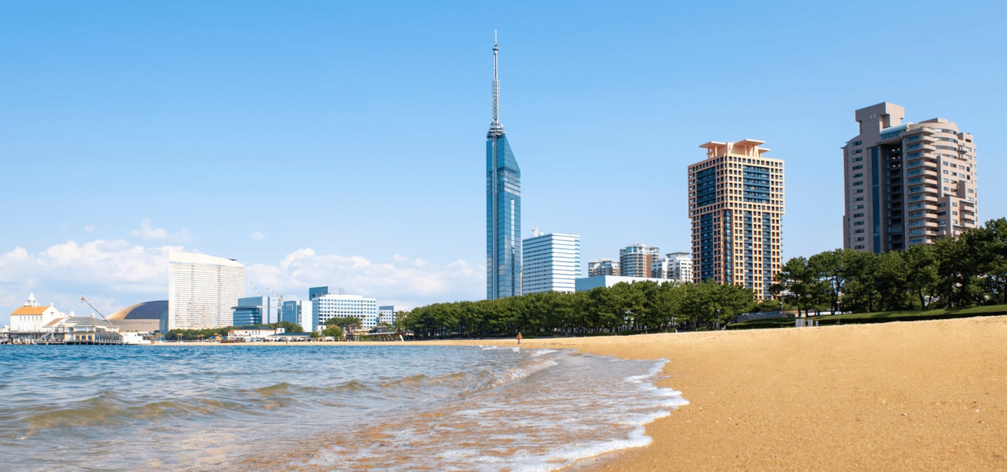 푸른 바다와 황금빛 모래사장이 펼쳐진 모모치 해변 너머로 우뚝 솟은 후쿠오카 타워와 세련된 도시의 스카이라인이 어우러진 모습입니다.