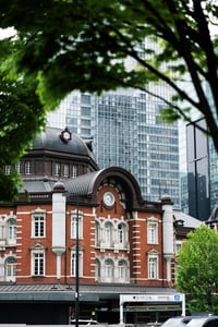 푸른 가로수 사이로 보이는 도쿄역의 붉은 벽돌 외관과 현대적인 고층 빌딩이 조화를 이루는 도심 속 풍경입니다.