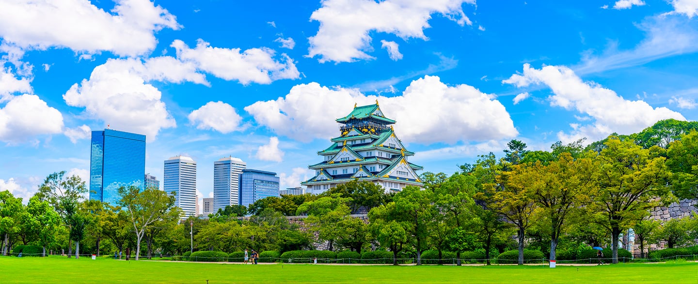 맑은 날씨 속 푸른 잔디밭 너머로 보이는 일본 오사카 성의 웅장한 천수각과 도심의 현대적인 빌딩 숲 전경입니다.