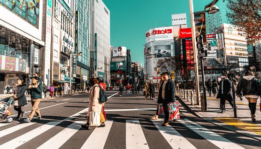 푸른 하늘 아래 신주쿠 도심의 횡단보도를 건너는 보행자들과 고층 빌딩의 화려한 간판들이 어우러진 일본의 활기찬 거리 풍경입니다.
