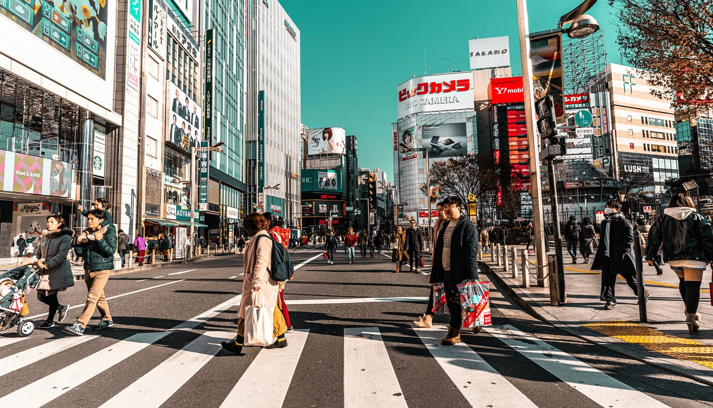 푸른 하늘 아래 신주쿠 도심의 횡단보도를 건너는 보행자들과 고층 빌딩의 화려한 간판들이 어우러진 일본의 활기찬 거리 풍경입니다.