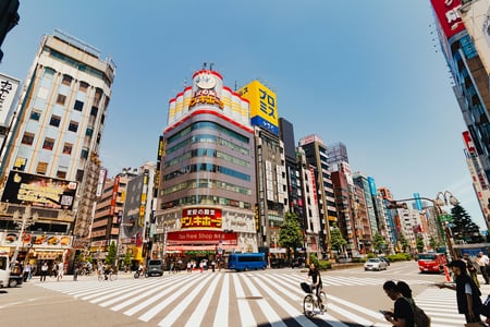 도쿄 신주쿠의 상징적인 돈키호테 건물과 넓은 횡단보도를 지나는 자전거 탄 사람과 행인들이 어우러진 활기찬 낮 시간대의 도심 전경입니다.