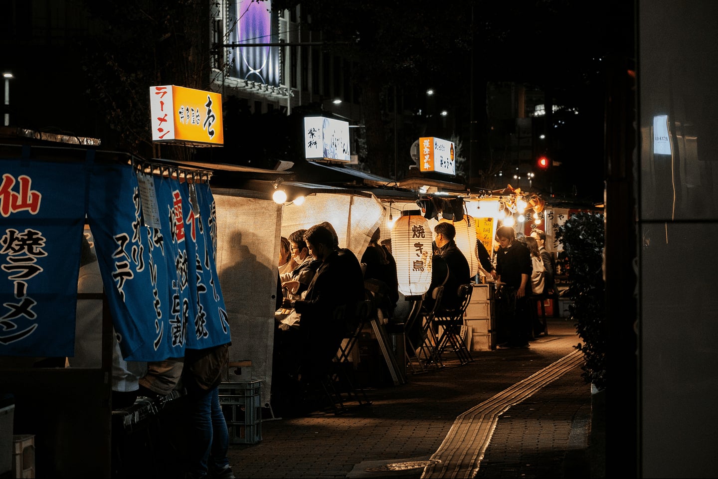 어두운 밤 일본 도심 거리에서 따뜻한 전등 불빛을 밝힌 포장마차 야타이에 사람들이 모여 앉아 식사를 즐기는 풍경입니다.