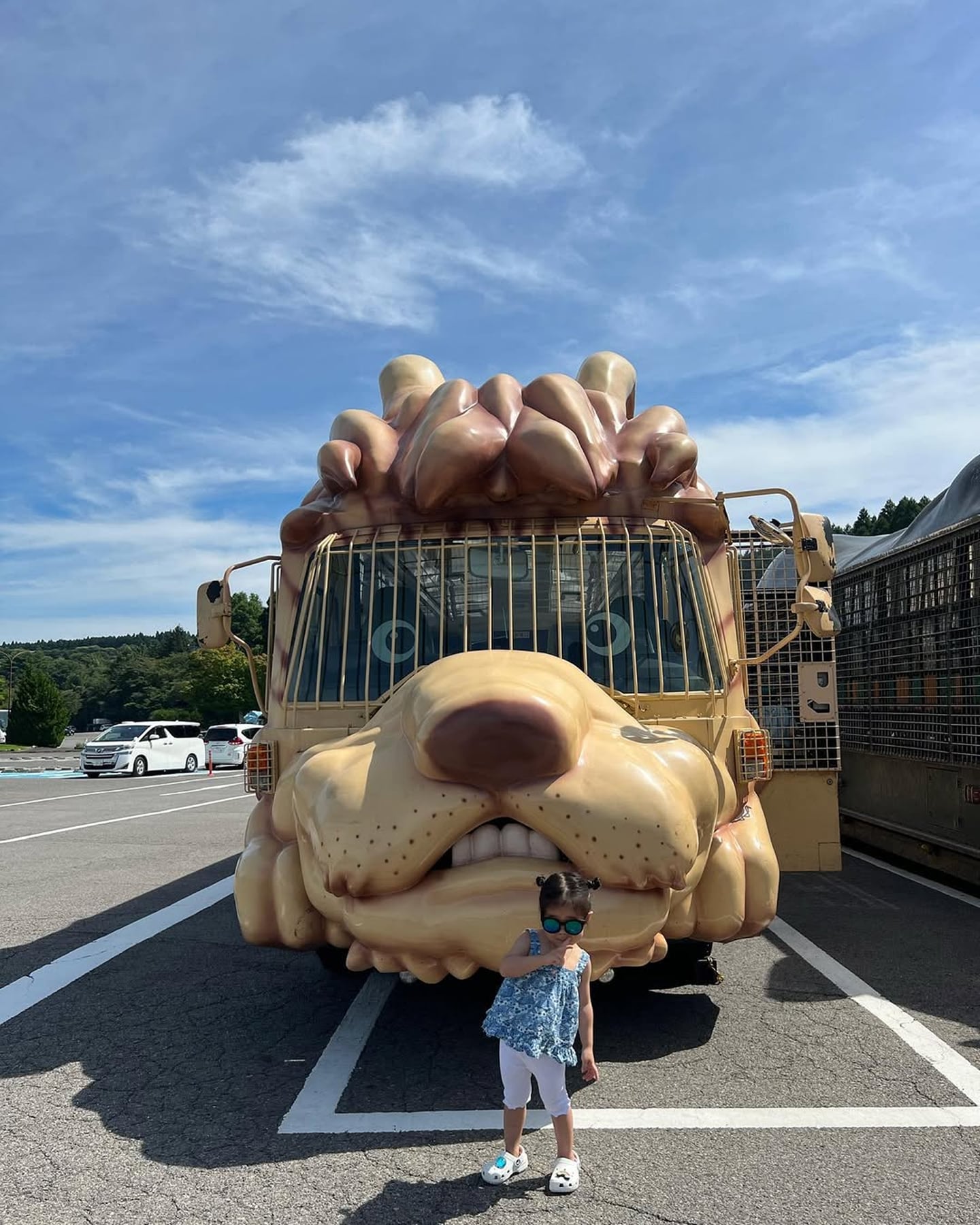 푸른 하늘 아래 주차장에서 거대한 사자 모양 사파리 버스를 배경으로 선글라스를 쓴 아이가 기념사진을 찍고 있는 모습입니다.