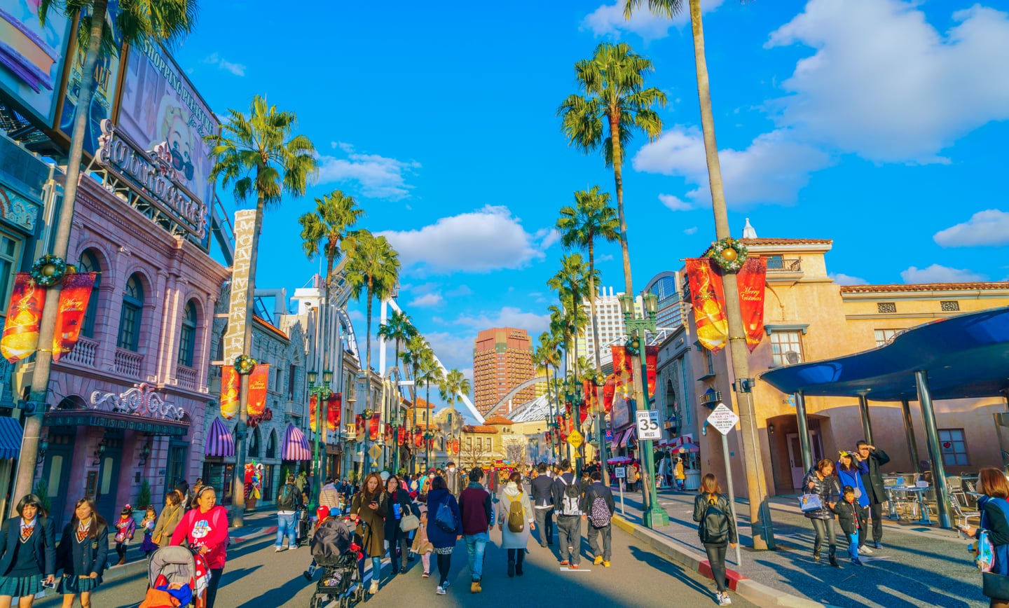 맑은 날씨의 테마파크 거리에서 야자수와 이국적인 건물들 사이로 인파가 붐비며 즐거운 시간을 보내는 모습입니다.