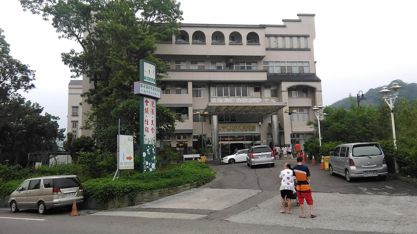 푸른 나무들에 둘러싸인 대만 관쯔링의 대형 온천 리조트 건물 앞으로 자동차와 사람들이 지나가는 평화로운 낮 풍경입니다.