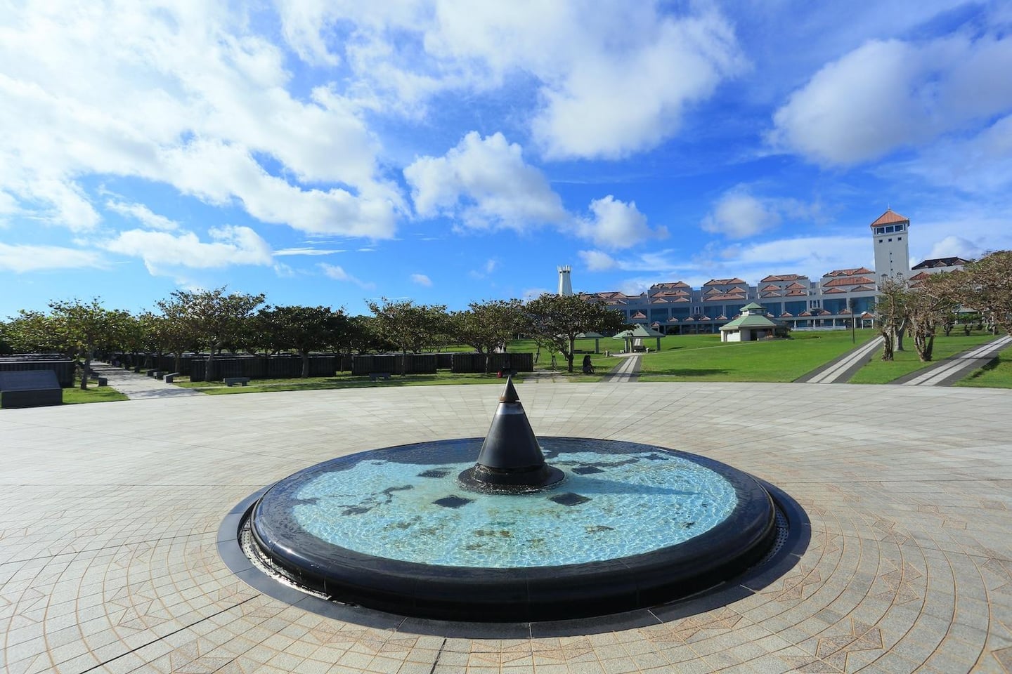 맑고 푸른 하늘 아래 원형 분수가 있는 넓은 석조 광장과 그 너머로 펼쳐진 푸른 잔디밭 및 리조트 건물이 조화를 이루는 풍경입니다.