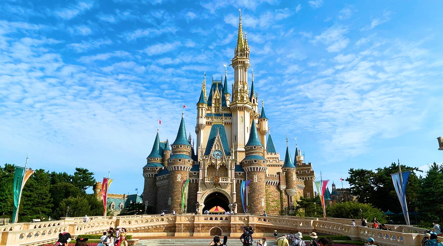 맑고 푸른 하늘을 배경으로 화려한 첨탑과 푸른 지붕이 돋보이는 거대한 성이 서 있고 앞쪽 광장에는 관광객들이 모여 있는 풍경입니다.