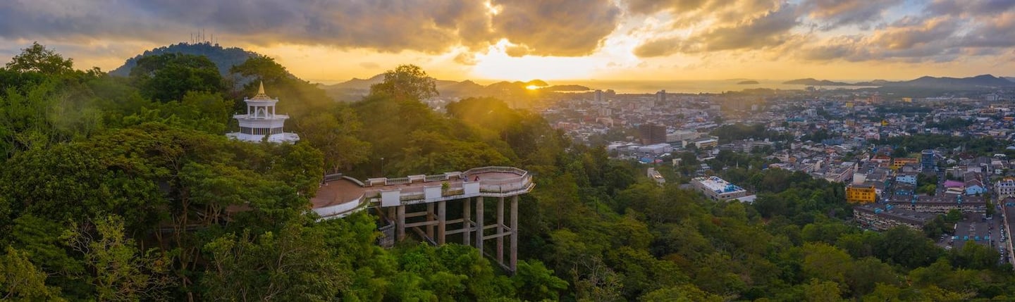황금빛 일몰이 내리쬐는 푸켓 카오랑 힐 전망대에서 바라본 울창한 숲과 평화로운 도시 전경이 파노라마처럼 펼쳐지는 풍경입니다.