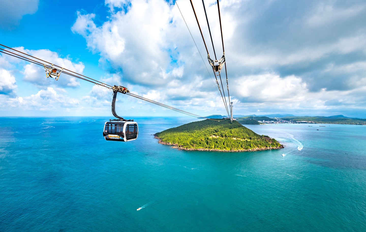 에메랄드빛 바다 위를 지나는 케이블카 너머로 초록빛 숲이 우거진 섬과 뭉게구름이 떠 있는 맑은 하늘이 펼쳐진 이국적인 풍경입니다.