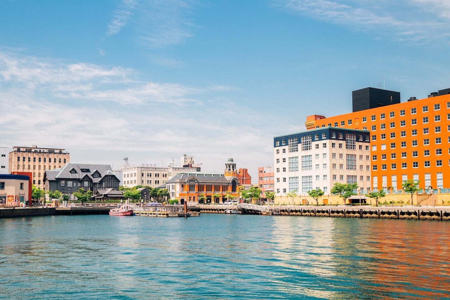 잔잔한 바다 물결 위로 주황색 대형 건물과 고풍스러운 유럽풍 건축물들이 늘어선 일본 모지코 레트로 항구의 맑고 화창한 풍경입니다.