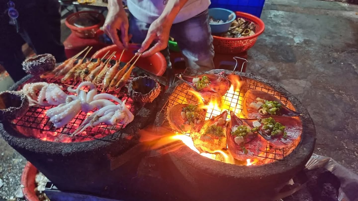 활활 타오르는 숯불 화로 위 석쇠에서 새우와 오징어, 양념된 조개를 정성스럽게 굽고 있는 베트남 야시장 길거리 음식 풍경입니다.