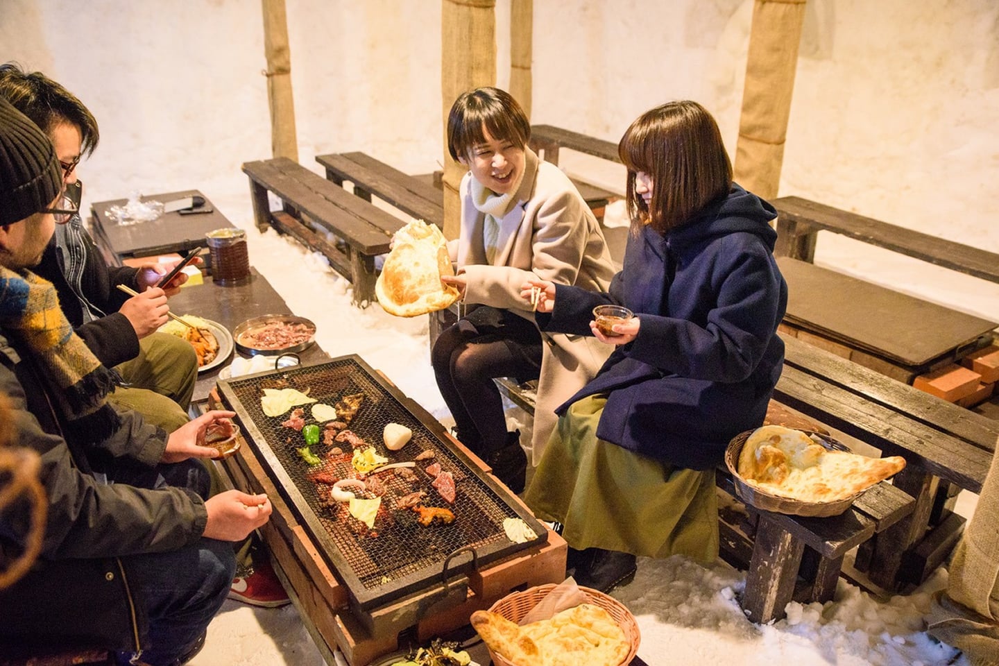 눈 덮인 겨울 야외 캠핑장에서 화로에 고기를 굽고 난을 곁들여 식사하며 즐겁게 대화하는 사람들의 모습입니다.