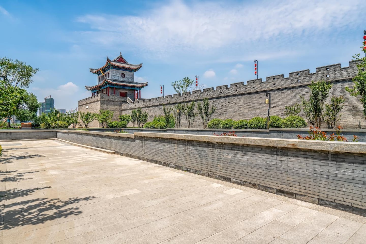 맑은 날씨 속에서 회색 벽돌로 정교하게 쌓인 고대 성벽과 화려한 전통 누각이 어우러진 풍경으로, 넓은 광장과 푸른 나무들이 평화로운 분위기를 자아냅니다.