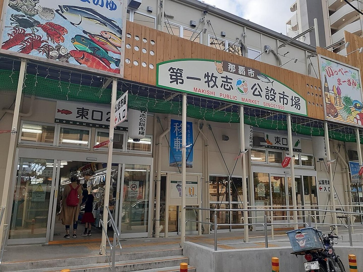 오키나와 나하시의 마키시 공설시장 입구로, 화려한 물고기 그림 간판과 일본어 안내판이 설치된 현대적인 시장 건물의 외관입니다.