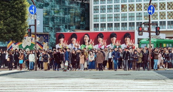 도쿄 시부야 스크램블 교차로 횡단보도 앞에 모여 신호를 기다리는 인파와 배경의 대형 인물 광고판이 어우러진 도심 풍경입니다.