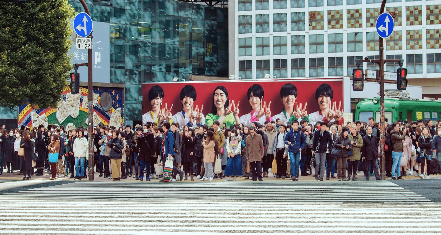 도쿄 시부야 스크램블 교차로 횡단보도 앞에 모여 신호를 기다리는 인파와 배경의 대형 인물 광고판이 어우러진 도심 풍경입니다.
