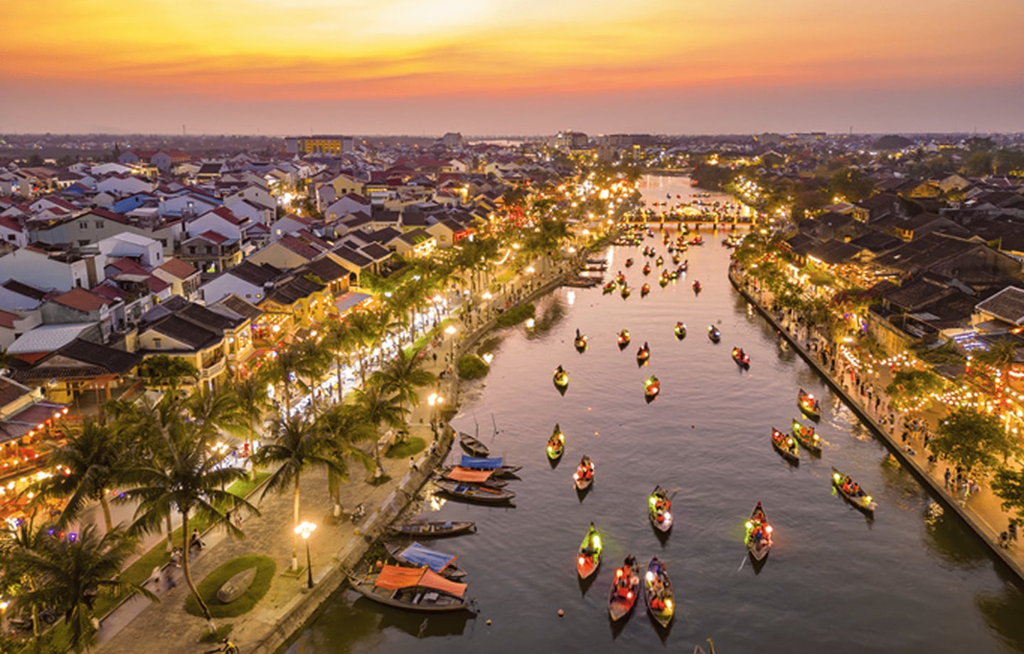황금빛 노을이 내리는 호이안 투본강 위로 등불을 밝힌 나룻배들이 줄지어 떠 있고 강변의 고풍스러운 건물들이 화려한 조명으로 빛나는 야경입니다.