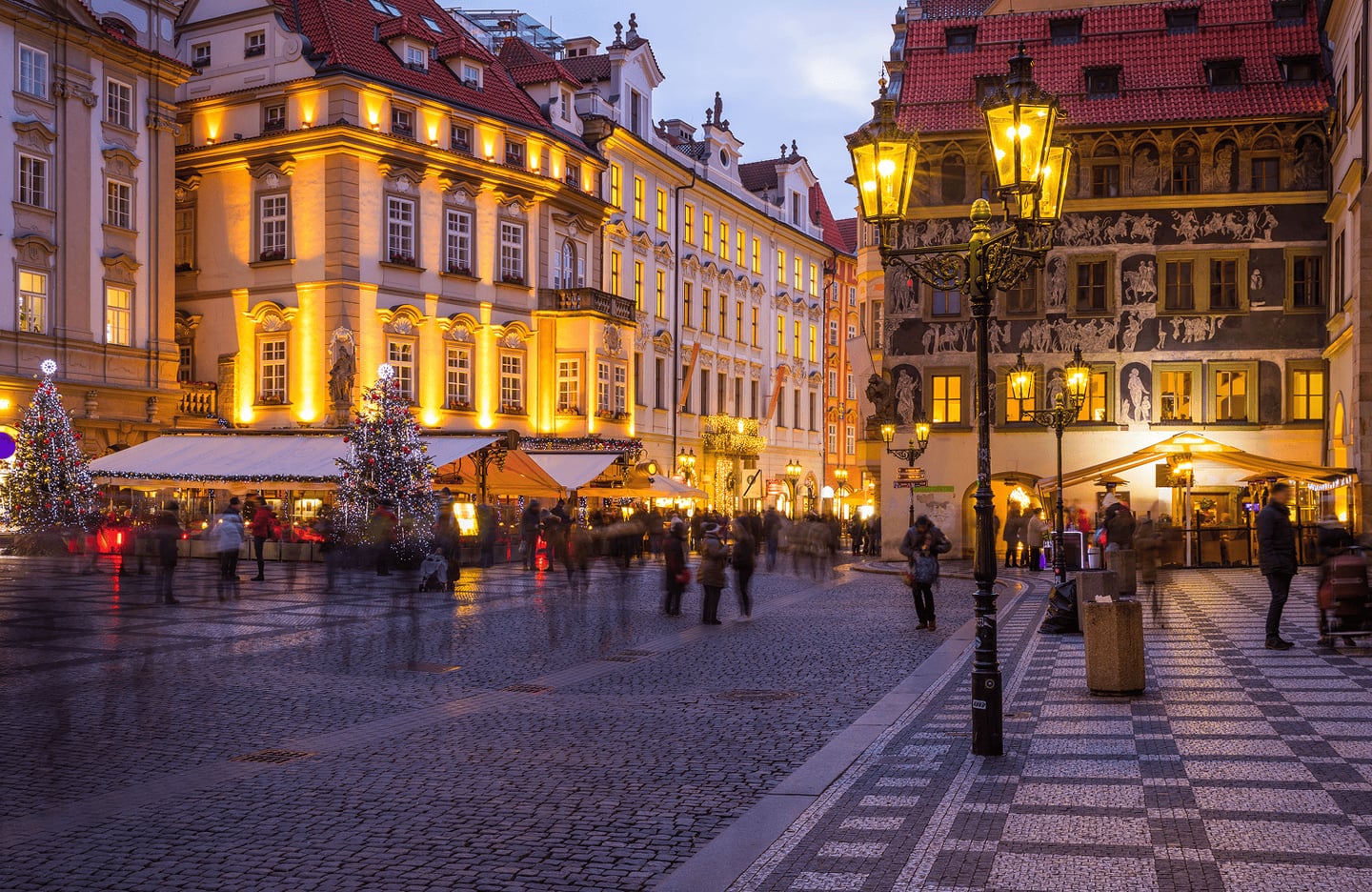 황금빛 조명이 켜진 유럽풍 건물들과 크리스마스트리가 장식된 프라하 구시가지 광장의 활기찬 저녁 풍경입니다.