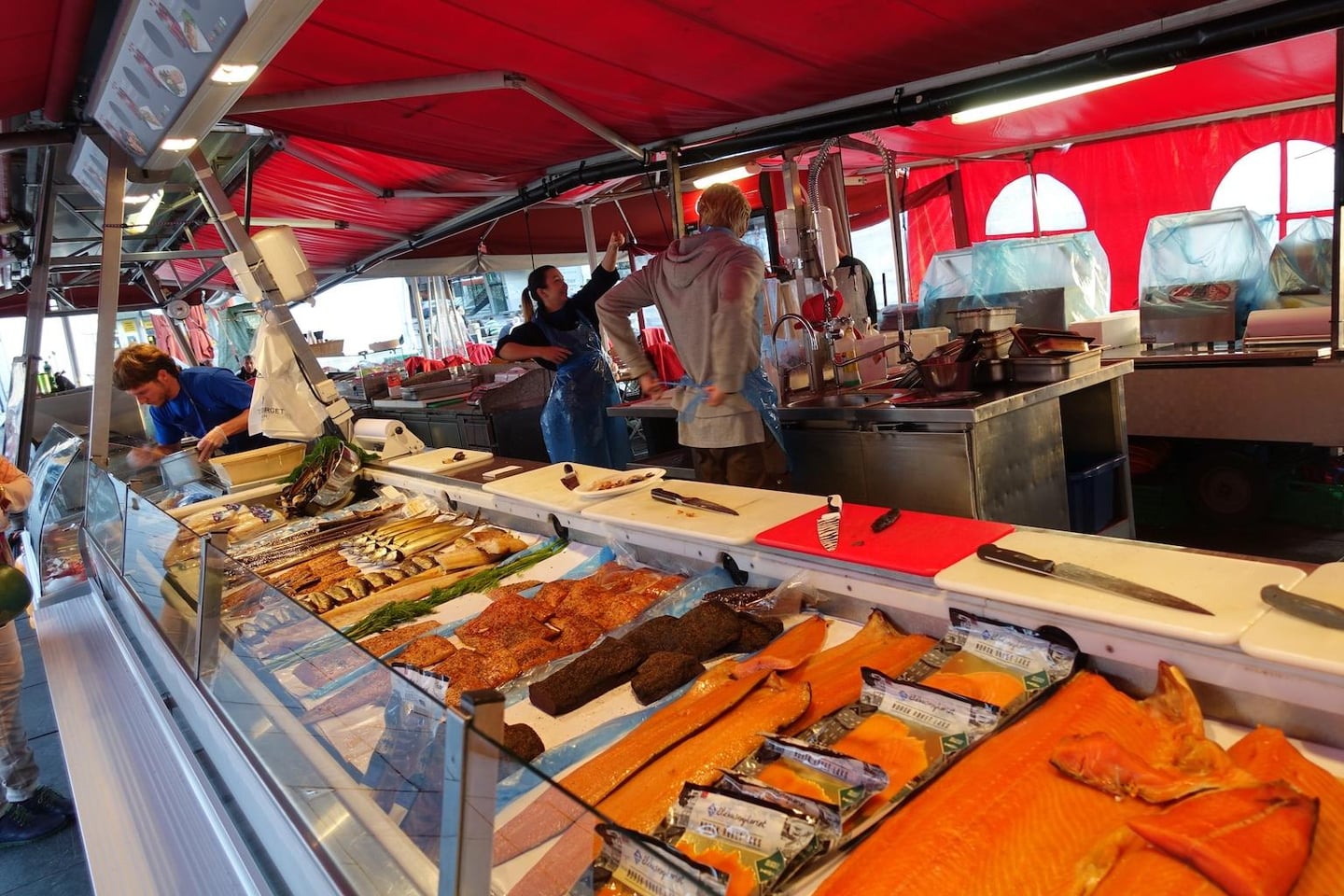 빨간 천막이 쳐진 야외 시장 매대 위에 먹음직스러운 주황빛 훈제 연어와 다양한 생선들이 진열되어 있고 상인들이 분주하게 움직이는 모습입니다.