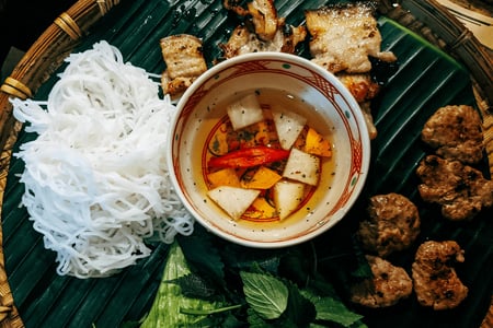 바나나 잎 접시 위에 놓인 하얀 쌀국수 면과 숯불 돼지고기 구이, 그리고 붉은 고추가 띄워진 느억맘 소스 그릇이 담긴 분짜 상차림입니다.