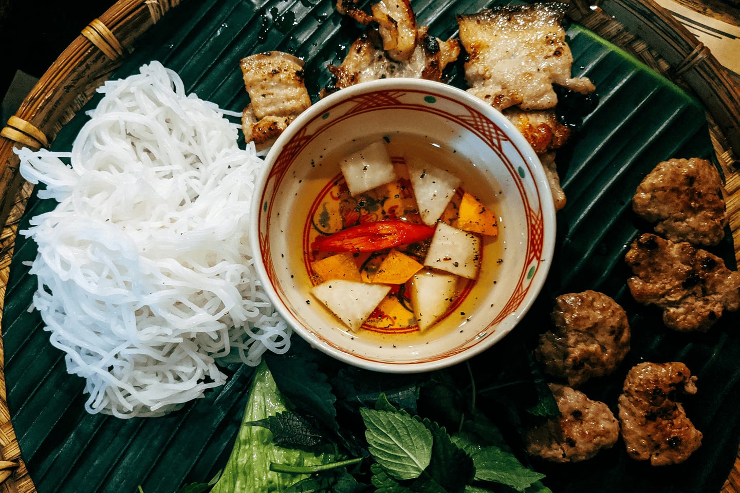 바나나 잎 접시 위에 놓인 하얀 쌀국수 면과 숯불 돼지고기 구이, 그리고 붉은 고추가 띄워진 느억맘 소스 그릇이 담긴 분짜 상차림입니다.