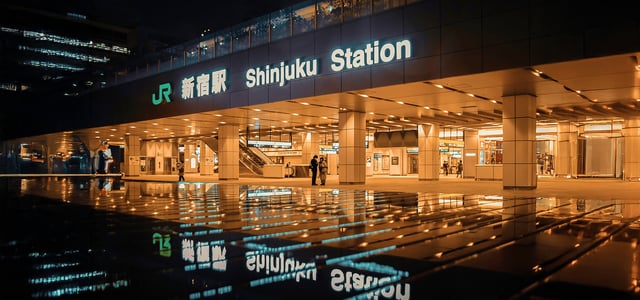 화려한 조명이 켜진 밤의 신주쿠역 입구 모습으로, 바닥에 반사된 역 이름 간판과 따뜻한 오렌지빛 조명이 어우러진 도쿄의 현대적인 야경입니다.