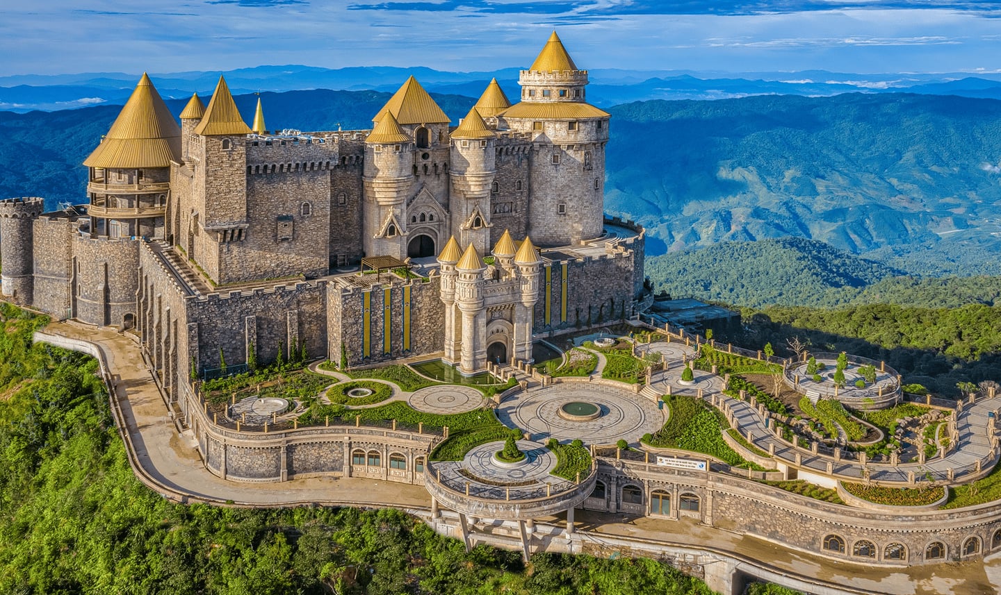 베트남 다낭 바나힐의 이국적인 풍경으로, 첩첩산중 속에 자리 잡은 황금빛 지붕의 거대한 석조 성과 기하학적 패턴의 정원이 조화를 이룹니다.