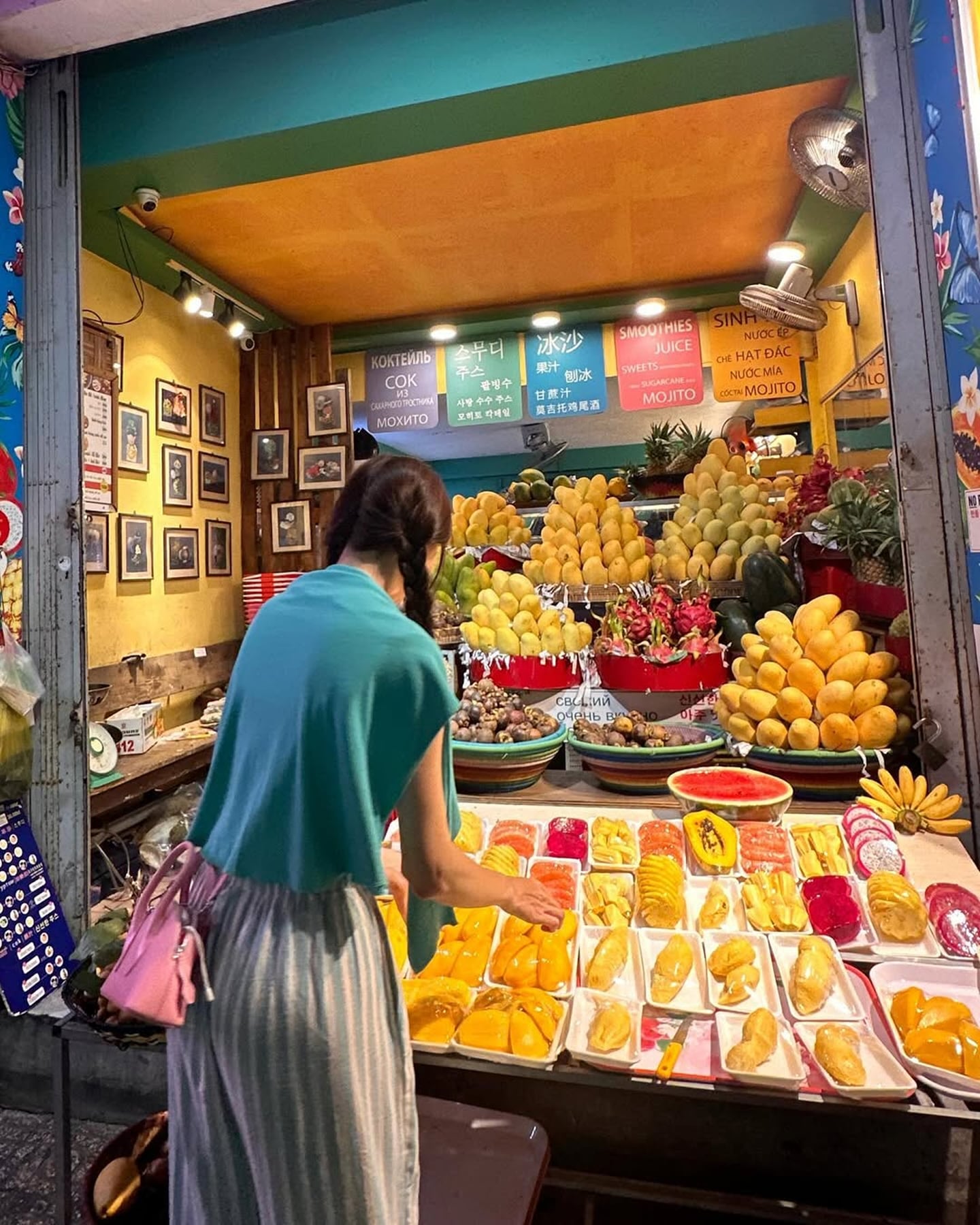 화려한 색감의 망고와 용과가 가득한 야시장 과일 가게에서 민트색 상의를 입은 여성이 과일을 고르고 있는 모습입니다.