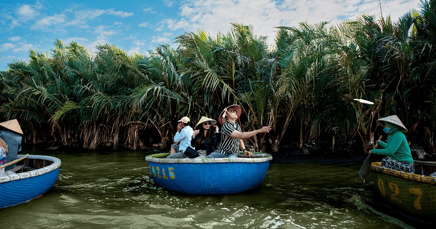 푸른 하늘 아래 울창한 야자수 숲을 배경으로 관광객들이 파란색 둥근 바구니 배를 타고 베트남의 전통적인 수로 여행을 즐기는 장면입니다.