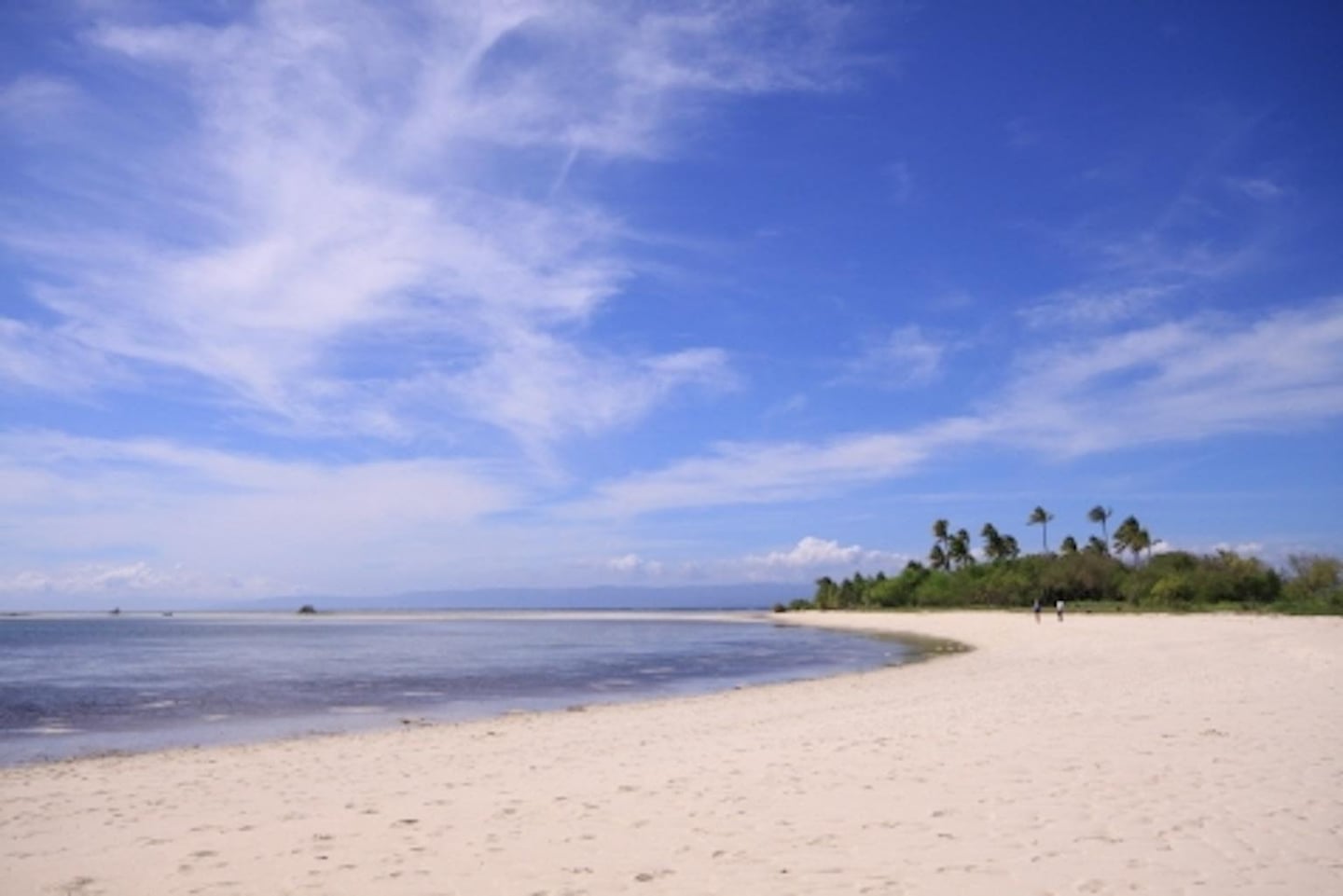 부드러운 곡선을 그리는 백사장과 투명한 바다 위로 깃털 같은 구름이 흩뿌려진 푸른 하늘이 펼쳐진 이국적인 해변의 전경입니다.