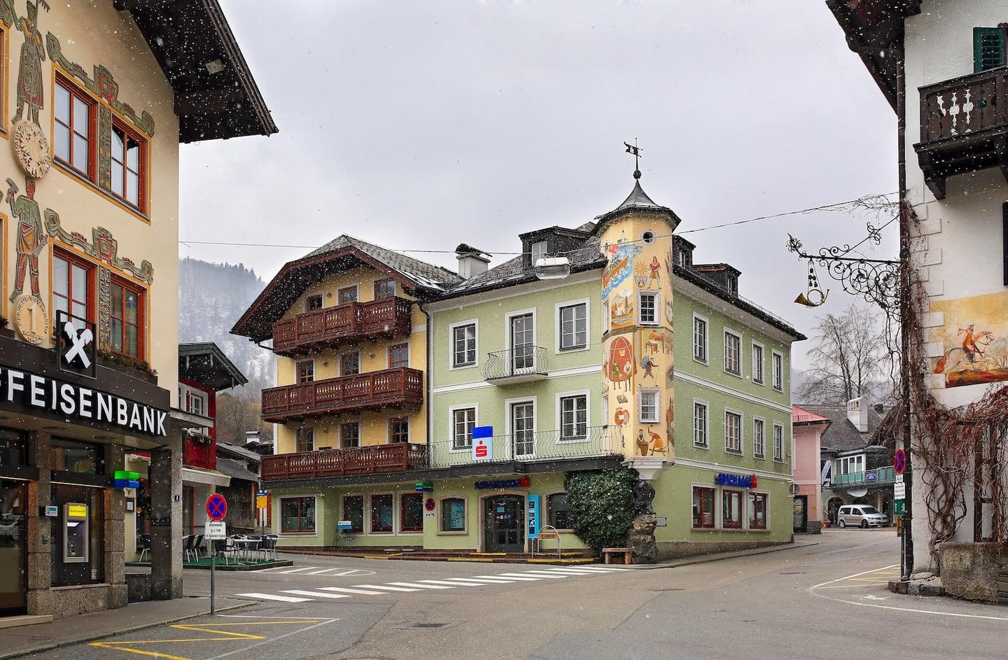 눈 내리는 겨울날, 벽화가 그려진 연두색 건물과 목재 발코니가 있는 전통 가옥들이 조화를 이루는 유럽 마을의 한적한 거리 풍경입니다.