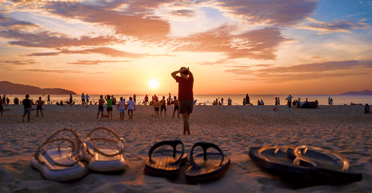 황금빛 노을이 지는 해변 모래사장 위에 놓인 샌들과 바다를 배경으로 여유로운 시간을 보내는 사람들의 실루엣이 어우러진 평화로운 풍경입니다.