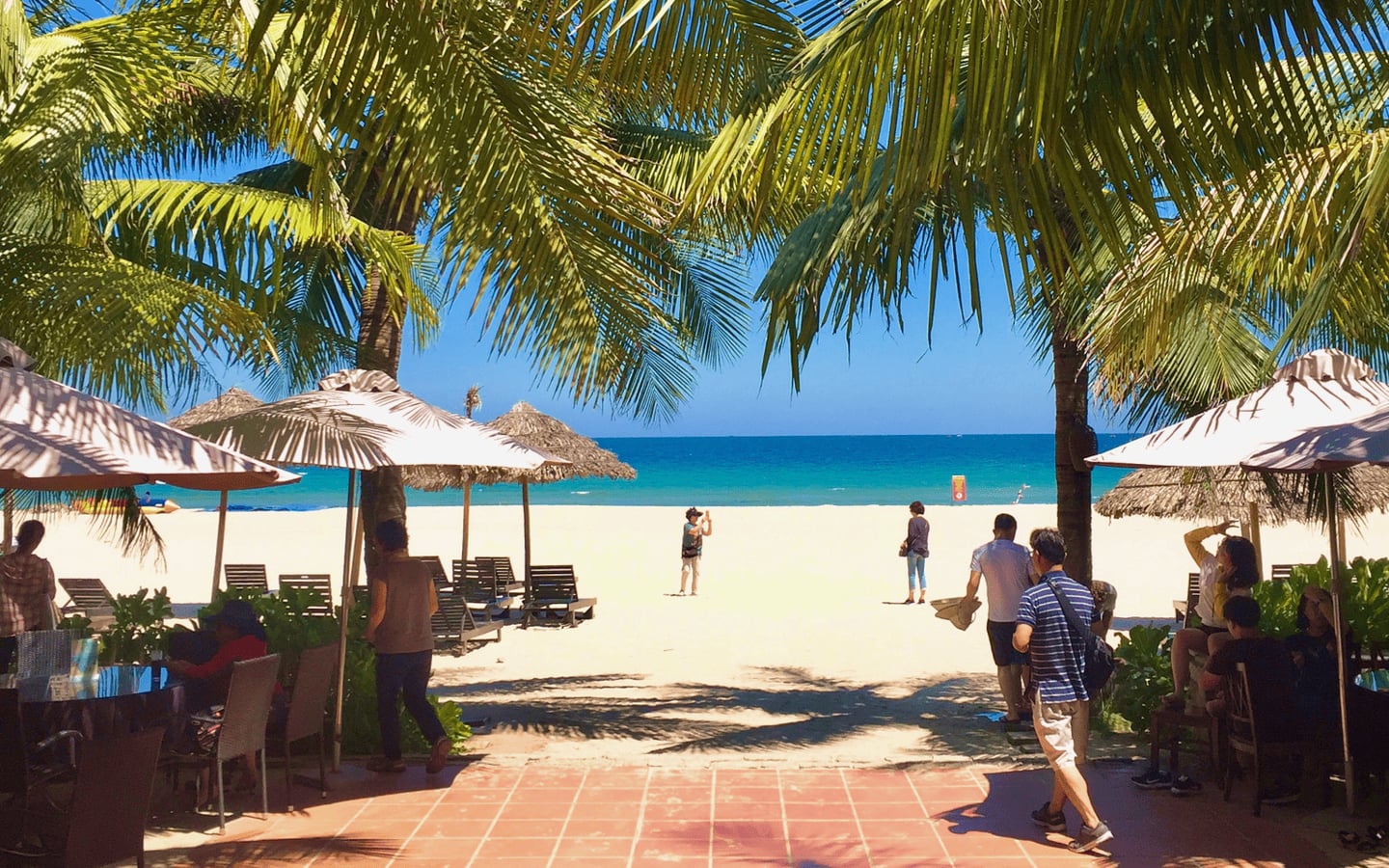 야자수 잎 사이로 보이는 에메랄드빛 바다와 하얀 백사장, 그리고 파라솔 아래에서 휴양을 즐기는 사람들의 여유로운 해변 모습입니다.