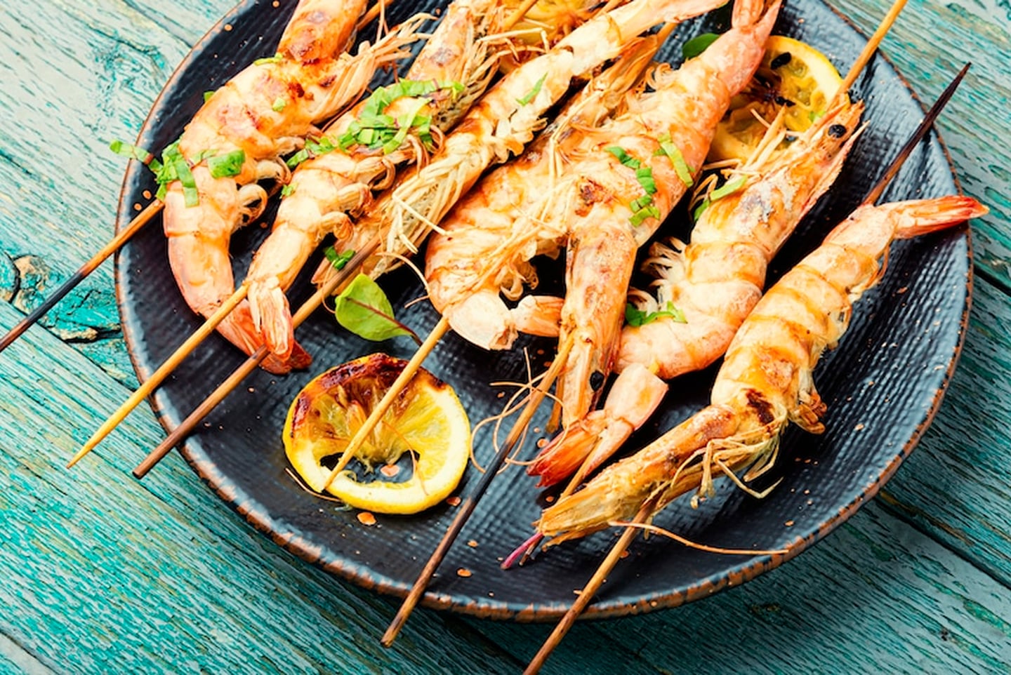 나무 꼬치에 꽂아 노릇하게 구운 새우 요리가 레몬 슬라이스와 함께 검은 접시에 정갈하게 담겨 있는 모습입니다.