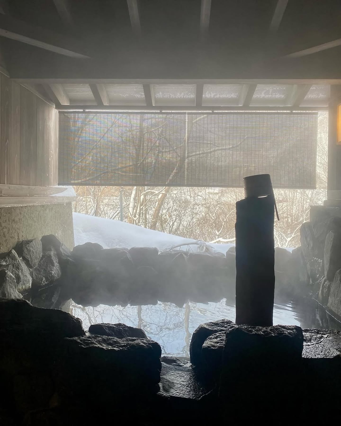 하얀 눈이 쌓인 숲을 배경으로 김이 모락모락 나는 따뜻한 노천 온천탕과 바위 주변에 놓인 나무 바가지가 어우러진 겨울 휴양지 풍경입니다.