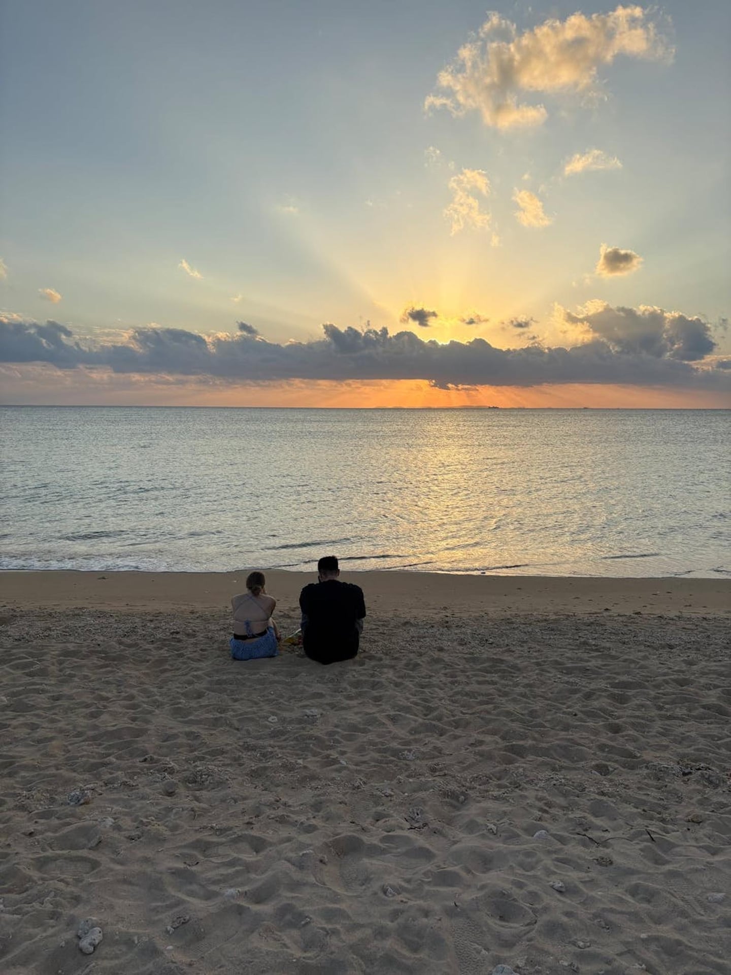 황금빛 노을이 지는 해변 모래사장에 앉아 바다를 바라보며 휴식을 취하는 커플의 뒷모습과 구름 사이로 비치는 햇살이 평화로운 분위기를 자아냅니다.