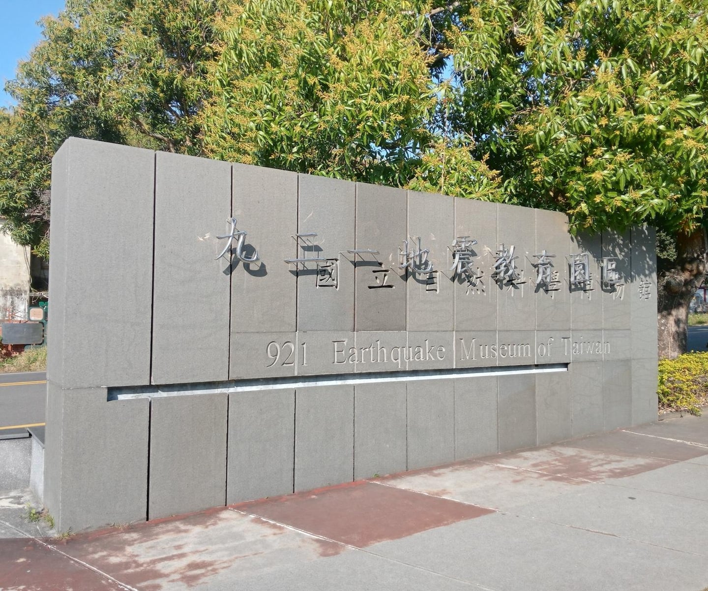 푸른 나무들이 배경으로 자리한 가운데 대만 921 지진 교육 박물관의 이름이 한자와 영어로 정갈하게 새겨진 회색 돌담 입구 전경입니다.